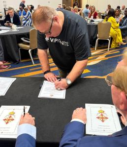 Michael Fomkin, wearing a VIP Ignite shirt, signs his official oath of knighthood for the Royal Order at a formal ceremony.