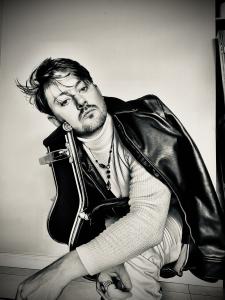 augustus caelthorne is displaying a vogue-like photo, of himself self-directed and self-photographed as part of his portfolio in monochrome colors wearing a black leather jacket, seated on a black leather chair