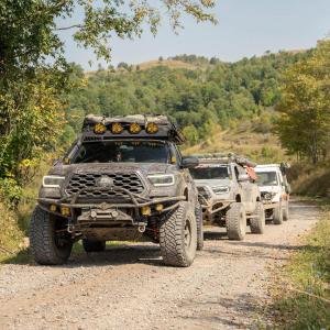 Toyota Tacoma with Ride Shocks leading off-road overland convoy on gravel trail through forested hills.