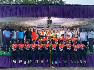 A women's sports team in matching uniforms sits in the front row, with coaches and officials standing behind them on a decorated stage outdoors.
