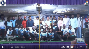 A men's sports team in matching uniforms sits in the front row, with coaches and officials standing behind them on a decorated stage outdoors.
