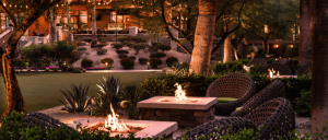 A serene outdoor lounge with glowing fire pits, surrounded by lush greenery and cozy seating, illuminated by warm evening lights.