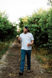 Ben Moore, founder of The Ugly Company, walks through a fruit orchard in California’s Central Valley, casually juggling a piece of fruit in one hand while holding another in the other hand. He wears a white Ugly Company t-shirt, jeans, and a backward cap,