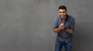 Smiling Sammer Affridi - Founder of HERO SHOT Photograpghy in casual grey shirt and jeans leans forward with clasped hands against a neutral grey studio backdrop—friendly personal-branding portrait.