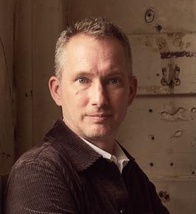 Portrait of Grant Gibson, one of the high-profile judges for this year’s Only Natural student design competition. He is seated against a rustic, textured background, wearing a brown corduroy jacket over a white shirt, and looking directly at the camera wi