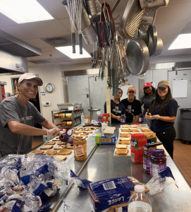 A group of women standing around a commercial kitchen table, preparing peanut butter and jelly sandwhiches.