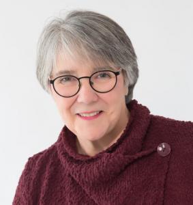 A smiling, humorous middle aged woman with short gray hair, laughing eyes, and chic black glasses