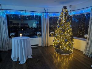 Interior holiday scene with a gold-lit Christmas tree decorated in blue and silver ornaments, surrounded by poinsettias, blue icicle lights, and a candlelit cocktail table near large windows overlooking a city skyline.