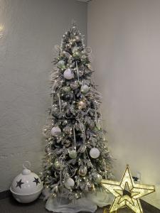 Elegant Christmas tree with silver and white ornaments, delicate ribbon, and frosted branches, placed next to a lit star decoration and a large white jingle bell with black stars in a softly textured corner of a room.