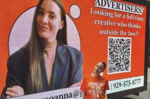 A woman standing next to an orange billboard truck with her face and a job pitch displayed, during a job-hunting stunt in New York City.