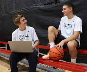 Two boys sitting on bleachers, smiling at each other. Both are wearing white tshirts and black shorts or pants. One has a laptop, and the other has a basketball in his hand.