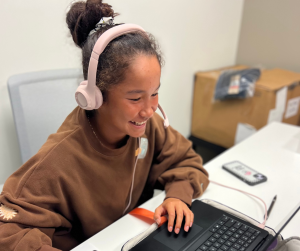 A student with tan skin, her hair in a bun. She has pink headphones on her head and is wearing a brown sweatshirt, seated at a desk studying.
