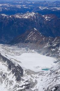Photo shows an aerial view of mountain peaks with a snow-covered glacier in the centre. A small opening in the ice showing green water is visible on right-hand side.
