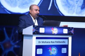 Dr. Mohana Rao Patibandla speaking at the podium during YNFNSICON, presenting on minimally invasive neurosurgery, with neuroimaging visuals in the background.