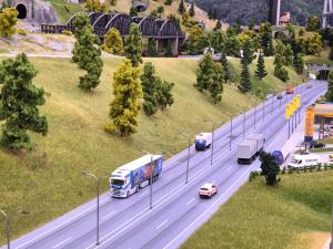 A realistic scale-model highway layout showing multiple lanes of traffic with trucks and cars passing a JET service station, surrounded by grassy embankments, streetlights, trees, and a railway bridge overhead.