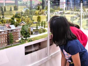 Children looking at a glass enclosed model railroad diorama featuring miniature houses, trees, winding tracks, and tiny figures in a detailed landscape.