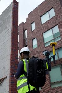 Female Surveyor using an Exyn Nexys portable SLAM mapping unit integrated with a Trimble DA2 GNSS unit at an urban construction site.