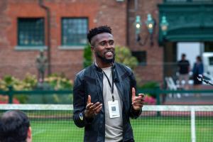 A person wearing a leather jacket and conference badge speaks to a group outdoors, using hand gestures and a microphone headset, with a tennis court and historic building in the background.