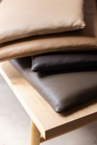 Four leather bench cushions in a variety of brown and tan colors, stacked messily on one another on top of a maple wood bench. Close up of corners of cushions to see the detail of the leather.