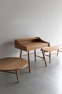 A lineup of wooden furniture in a bare room with cement floors and white walls. From left to right: round Hudson Coffee Table, new Hudson Desk, Hudson Desk