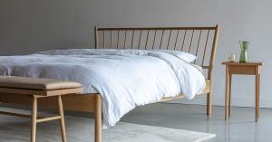 Solid white oak wood bedroom furniture including a spindle bed with white bedding, a turned leg bench with a light tan leather cushion, and a simple side table with a green class cup and pitcher of water