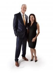 Image of Bob and Catherine Hilliard. Bob is a tall gentleman, with white hair, wearing a dark suit and yellow tie. Catherine is a hispanic woman with long brown hair. She wears a sleeveless black dress.