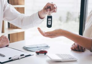 A person hands over car keys to a new owner across a desk with money, a contract, and a toy car—symbolizing the excitement of ownership and the hidden financial responsibilities that come with it.
