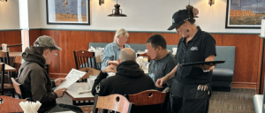 A waitress assists a group of diners at a cozy restaurant table, with menus and condiments visible. Brightly lit and inviting atmosphere.