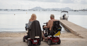 Elderly couple riding 4-wheel portable electric mobility scooters near the ocean, enjoying summer independence and outdoor exploration.