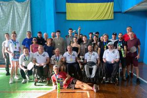 Large group of Ukrainian pickleball players and organizers—including adaptive athletes—pose indoors under a Ukrainian flag, holding medals, trophies, and paddles.
