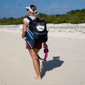 Woman walking barefoot on beach carrying fins and BLU3 Nomad tankless dive system in branded backpack for portable diving.