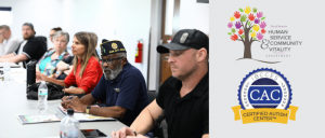 A group of individuals seated at a table during a community meeting, with logos for a human services department and certified autism center.