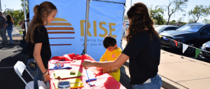 Children and adults engage in a colorful arts and crafts activity at a RISE event, with a vibrant banner in the background.
