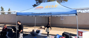A colorful tent with "RISE" branding shelters people engaging in activities, with toys and materials scattered on the ground.