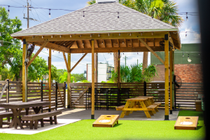 Outdoor seating area at The Yardery featuring picnic tables under a wooden pavilion, surrounded by yard games including two cornhole boards on artificial turf.