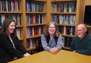 Two women and a man sit smiling at a conference table. There are bookshelves behind them.