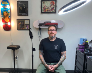 Tattoo artist Jason “Elvis” Martin seated in his studio, smiling, with framed art and tattoo supplies visible in the background.