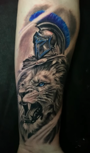 Realistic tattoo of a roaring lion wearing a Spartan warrior helmet with blue plume accents, inked in black, grey, and blue.