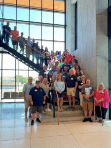 A large group of approximately 30-40 people posed together on and around a modern staircase in a bright, contemporary building with floor-to-ceiling windows and stone walls. The group includes men and women of various ages, many wearing name tags and casu