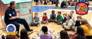 A firefighter sits on a chair in a gym, teaching a group of children gathered around him, with playhouses in the background.