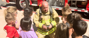 A firefighter in full gear sits on the ground, engaging with a group of children who are attentively listening and curious.