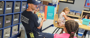 A firefighter reads a colorful children’s book to captivated young girls in a classroom setting, fostering a love for reading.