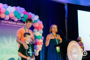 Woman, man, and boy--wearing conference lanyards--stand on stage at an NSA Annual Conference. The stage is decorated with a balloon arch and purple and turquoise backdrop.
