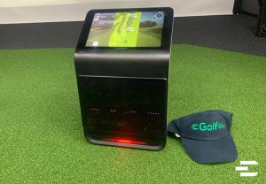 Garmin Approach R50 launch monitor displaying a virtual golf course, shown on green turf with a Golfible branded hat beside the device
