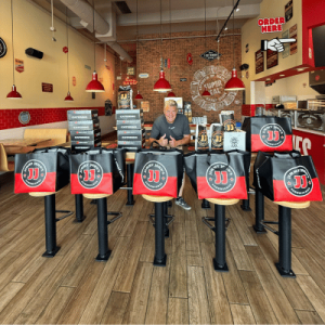 A man sits in a sandwich shop surrounded by large bags with "JJ" logos, preparing for orders against a cozy, red-themed interior.