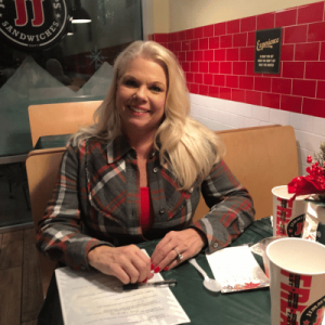A woman in a plaid shirt sits at a table in a sandwich shop, holding a pen and a menu, surrounded by festive decor.