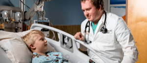 A doctor in a white coat examines a child wearing a hospital gown in a medical room, discussing the child's condition.