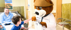 Mettu Children's Hospital's friendly bear mascot in a doctor's coat interacts with a young boy in a hospital room, promoting a cheerful atmosphere.