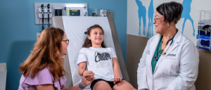 A young girl in a hospital gown sits on an exam table, while two adult women, one in a lab coat, discuss her care.