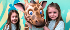Two children in matching light blue shirts pose with a large, animated giraffe head against a colorful, playful background.